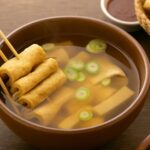 Korean fish cake soup served in a traditional bowl with green onions and skewers